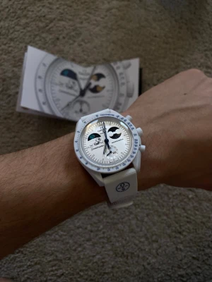 Omega x Swatch MoonSwatch Moonshine Snow - Säljer en Omega x Swatch MoonSwatch Mission to Earthphase Moonshine Gold Snoopy Snowflake winter exclusive med vit boett och armband. Klockan har detaljer i svart och blått, månfaskomplication och biokeramisk konstruktion. Snygg och unik design med tydliga visare och index. Levereras i originalbox.