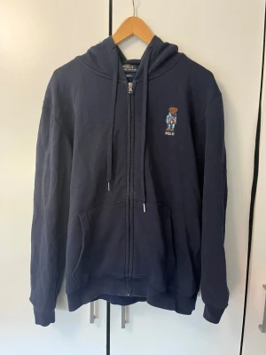 Mörkblå hoodie från Polo Ralph Lauren - Snygg mörkblå hoodie från Polo Ralph Lauren med dragkedja, huva och fickor framtill. På bröstet finns den klassiska björnen broderad. Tillverkad i mjukt bomullsmaterial och har en relaxed fit som passar perfekt till vardagslooken.