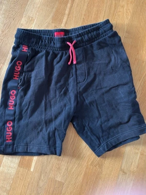 Svarta shorts från HUGO med röd logga - Snygga svarta shorts från HUGO med röda detaljer och logga längs sidorna. Shortsen har elastisk midja med röd snörning och är tillverkade i mjuk bomull. 