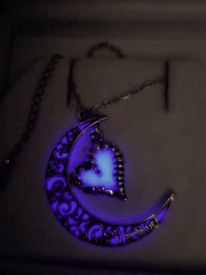 Halsband med måne och lysande hjärta - Unikt halsband i silverfärgad metall med en halvmåne dekorerad med snirkliga mönster och ett hängande hjärta som lyser lila i mörkret efter att ha absorberat dagsljus eller solljus en kort stund. Kedjan är tunn i rostfritt stål och har justerbar längd. Perfekt för dig som gillar magiska detaljer och vill sticka ut. Både hänget & kedjan tål vatten utan att missfärgas & har en lång livslängd. Hänget är ca 3 cm och kedjan 50 cm.