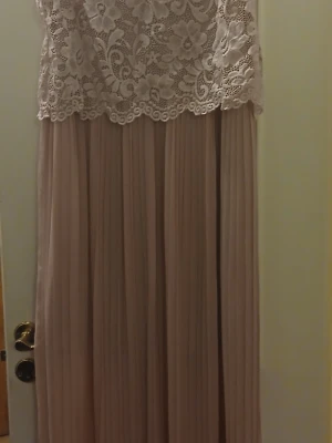 Lång spetsklänning från Happy Holly - Elegant långklänning från Happy Holly i ljus beige ton. Överdelen har vacker blommig spets och rund halsringning, medan kjolen är plisserad och faller mjukt. Perfekt för speciella tillfällen där du vill känna dig extra fin.