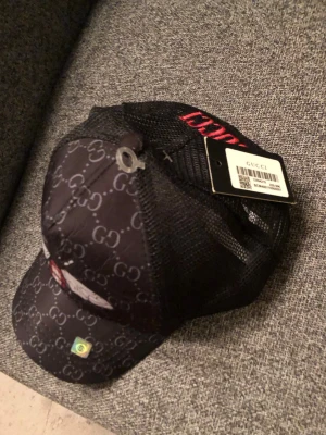 NY Snygg keps GG med Motiv - Snygg svart keps från Gucci med klassiskt GG-mönster över hela kepsen och mesh på baksidan för extra ventilation. Framtill finns en liten patch och detaljer i rött. Märkeslapp och etikett sitter kvar. Perfekt för dig som vill ha en exklusiv och trendig look.