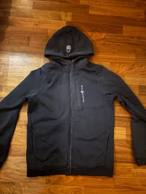 Svart hoodie från Sail Racing - Svart hoodie från Sail Racing med dragkedja framtill och logga på bröstet samt huvan. Tillverkad i mjukt material, perfekt för en avslappnad och sportig stil. Snygga detaljer och bekväm passform.