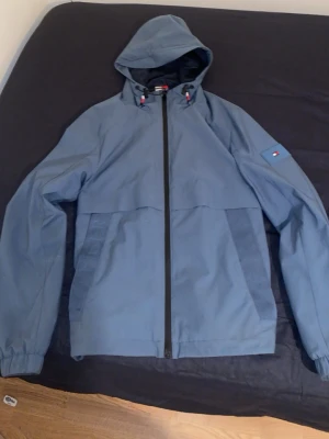 Blå vindjacka från Tommy Hilfiger - Snygg blå vindjacka från Tommy Hilfiger med huva och hel dragkedja framtill. Jackan har diskret logga på ärmen och 'HILFIGER' tryckt på ryggen. Praktiska sidofickor och elastiska muddar vid ärmsluten. Perfekt för blåsiga dagar och enkel att matcha. Pris går p diskutera desto snabbare man köper varan extrem snygg jacka knappt använd som ny dyr men säljer billigt och bra passform den passar m 