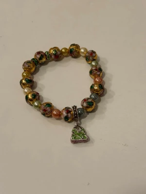 Guldtonat armband med väsk-charm - Unikt armband med pärlor i guld, grönt, rosa och vitt samt en berlock formad som en grön handväska. Pärlorna har blommönster och metallicfinish. 