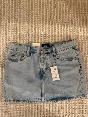 Ljusblå jeanskjol från 157 - Säljer en ljusblå jeanskjol från 157, modell Maddy. Kjolen har klassisk femficksdesign, bälteshällor och knappgylf. Perfekt basplagg till garderoben och enkel att styla med olika toppar.