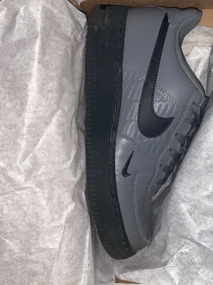 Nike Air Force 1 grå/svarta sneakers - Nike Air Force 1 sneakers i grått med svart swoosh och svart sula. Klassisk låg modell med ovandel i läder och snygga detaljer längs sidan. Perfekta för dig som vill ha en clean och stilren look med streetkänsla.