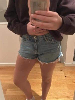Blå jeansshorts med fransig kant - Säljer ett par klassiska blå jeansshorts med fransig, rå kant och femficksmodell. Shortsen har hög midja och stängs med knapp och dragkedja. Perfekta för sommaren och lätta att matcha med allt.