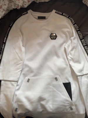 Vit hoodie från Philipp Plein - Säljer en vit hoodie från Philipp Plein med svarta detaljer och logga på bröstet. Svarta band med text längs ärmarna och stor magficka framtill. Materialet är mjukt och skönt, perfekt för en streetwear-look. Snygg detalj med mesh på fickan och tryck på ryggen.