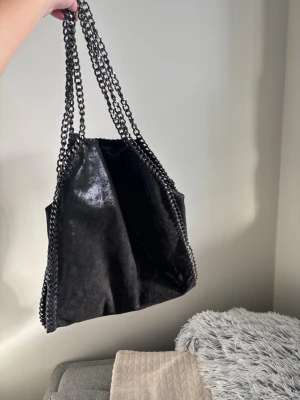 Svart axelväska med kedjedetaljer - Snygg svart axelväska i mjukt material och  silverfärgade kedjor som axelband och runt kanterna. Ger en cool, trendig vibe. Perfekt för dig som gillar statement-accessoarer.