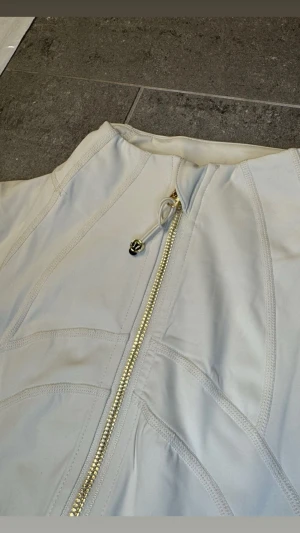  Vit Lululemon Define Jacket -                                                    Snygg och stilren träningsjacka från Lululemon i klassiskt svart med exklusiva guldfärgade dragkedjor som ger en lyxig touch ✨ Jackan är figurnära med smarta sömmar som framhäver silhuetten på ett väldigt smickrande sätt.  Perfekt för träning, yoga, gym eller som en sportig vardagsjacka. Materialet är stretchigt, följsamt och bekvämt – precis den höga kvalitet som Lululemon är kända för. Två praktiska fickor med drag