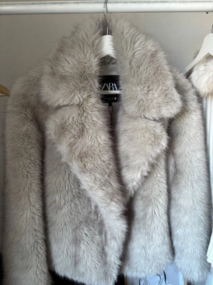 Beige fuskpälsjacka från Zara - Snygg beige fuskpälsjacka från Zara med bred krage och klassisk rak passform. Jackan har en lyxig känsla och är perfekt för att hålla värmen under kalla dagar. Mjuk och fluffig yta som ger en trendig look.