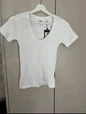 Vit v-ringad t-shirt från 157 - Klassisk vit t-shirt från 157 med v-ringning och korta ärmar. Tillverkad i mjuk bomull som känns skön mot huden. Enkel och stilren design som passar till allt. Perfekt basplagg för garderoben.