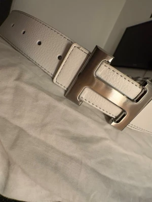 Vitt skärp med stor H-spänne - Snyggt vitt bälte med en rejäl H-formad metallspänne. Bältet har en stilren och modern look med synliga sömmar och är gjort i ett slätt material som liknar skinn. Perfekt för att lyfta vilken outfit som helst.