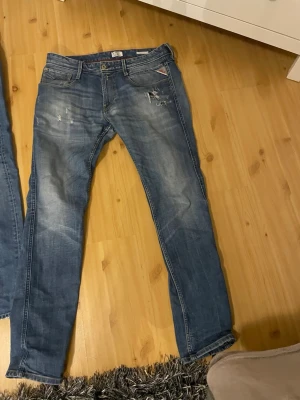 Blå slitna jeans från replay - Säljer ett par blå jeans från Diesel med slitningar och lätt tvättad look. Klassisk femficksmodell med raka ben och låg midja. Jeansen har detaljerade slitningar framtill och en liten röd Diesel-logga vid fickan. Perfekta för en avslappnad stil.