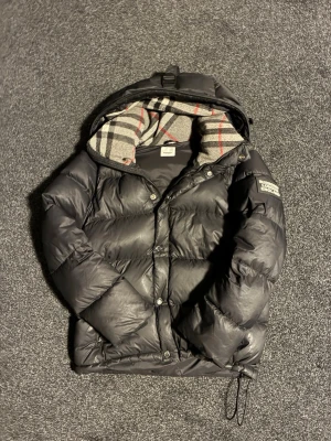 Burberry jacka - Säljer en svart pufferjacka från Burberry med klassiskt rutigt foder i huvan. Jackan har dragkedja, tryckknappar och justerbar nederkant. Puffig modell med stora kanaler och två sidofickor. Perfekt för kalla vinterdagar .