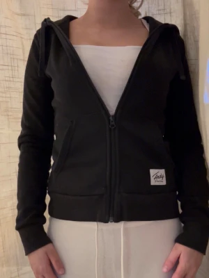 Svart hoodie från Andy med dragkedja - Säljer en svart hoodie från Andy med hel dragkedja framtill och två fickor. Hoodien har en klassisk passform, långa ärmar och en liten logotyp på vänster sida. Perfekt för en chill och avslappnad stil.