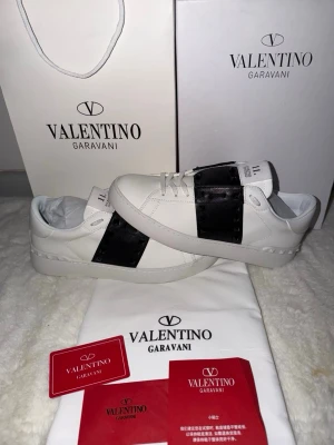 Valentino Garavani vita sneakers - Snygga vita sneakers från Valentino Garavani med en bred svart läderdetalj över sidan. Klassisk låg modell med vita snören och diskret logga på plösen. Skorna har en stilren design och är tillverkade i läder för en lyxig känsla.