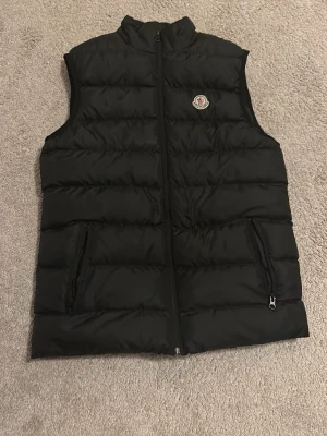 Svart dunväst från Moncler - Snygg svart dunväst från Moncler med hög krage och dragkedja framtill. Västen har två fickor med dragkedja och det klassiska Moncler-logotypen på bröstet. Perfekt för lager-på-lager och riktigt clean look.