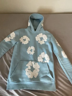 Ljusblå Denim tears hoodie med molntryck - Säljer en ljusblå hoodie med stora vita molntryck över hela plagget. Hoodien har en klassisk huva och en stor magficka framtill. Tillverkad i mjukt bomullsliknande material och har ribbade muddar vid ärmslut och nederkant. Perfekt för dig som gillar unika och lekfulla plagg. (bästa kopian du kan hitta) (pris kan diskuteras)