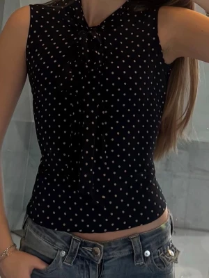 Svart prickig blus med knyt - Svart ärmlös blus med vita prickar. Figurnära passform som ger en chic känsla. Perfekt att styla med jeans eller kjol för en trendig look.
