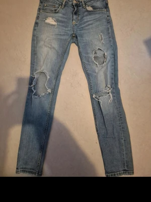 Blå slitna jeans från gina tricot - Säljer ett par ljusblå skinny jeans från ,gina tricot modell KRISTEN. Jeansen har flera stora slitningar och hål på benen för en trendig, avslappnad look. Klassisk femficksmodell med dragkedja och knapp. Perfekta för dig som gillar streetstyle sopig vibe.