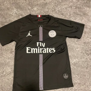 Svart PSG x Jordan matchtröja - Snygg svart matchtröja från PSG i samarbete med Jordan. Tröjan har vita loggor, Fly Emirates-tryck på bröstet och ett diskret randigt mönster i mitten. Tillverkad i lätt och ventilerande material, perfekt för fotboll eller träning.