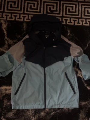 Nike Miller  - Snygg vindjacka från Nike i ljusblått, mörkblått och grått. Jackan har huva, dragkedja framtill och två fickor med dragkedja. Perfekt för blåsiga dagar och sportiga outfits. Tillverkad i lätt och tåligt material som står emot vind.