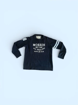  Morris tröja - Snygg marinblå stickad tröja från Morris med vita tryckdetaljer på bröstet och ärmen. Tröjan har rund hals, ribbade muddar och två vita ränder på ena ärmen. Perfekt för dig som gillar klassisk college-stil.