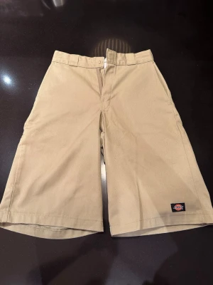 Beige shorts från Dickies - Klassiska beige chino shorts från Dickies med rak passform och breda ben. Shortsen har hällor för bälte, sidofickor och bakfickor. Tillverkade i slitstarkt material med Dickies-logga på benet. Perfekta för en chill och avslappnad stil. Passar S och M
