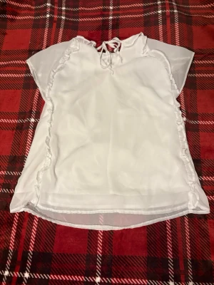Vit transparent blus med volangdetaljer - Söt vit blus i transparent material med volangdetaljer längs sidorna och knytning i halsen. Blusen har korta ärmar och v-ringad hals..