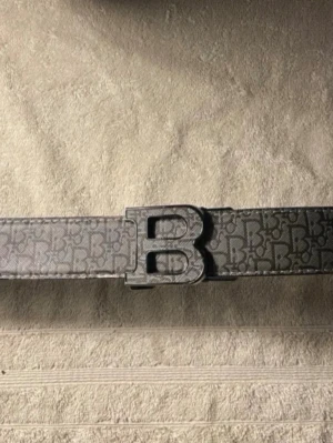 Svart mönstrat bälte med B-spänne - Snyggt svart bälte från Balmain med ett stort B-format metallspänne. Bältet har ett diskret mönster med små B-loggor över hela bandet och är gjort i skinn. Perfekt för att lyfta vilken outfit som helst.