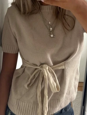 Beige stickad topp med knytband - Snygg beige stickad topp med korta ärmar och rund hals. Toppen har ett dekorativt knytband i midjan som ger en feminin siluett. Ribbstickade muddar vid ärmslut och nederkant. Perfekt att styla med både jeans och kjol. Intressekoll 