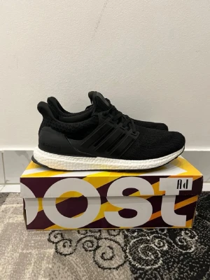 Adidas Ultraboost svarta sneakers - Svarta Adidas Ultraboost sneakers med stickad ovandel och klassiska tre ränder på sidan. Skorna har vit Boost-mellansula för extra komfort och svart yttersula. Snygg och sportig design med snörning och diskret logga på hälen. Perfekta för dig som gillar streetwear och sportig stil.