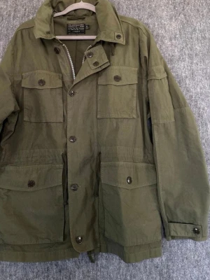 Olivgrön militärinspirerad jacka - Snygg olivgrön jacka från Alpha Industries med fyra stora fickor framtill, knappar och dragkedja. Klassisk militärstil med hög krage och justerbar midja. Perfekt för dig som gillar streetwear och utility-look.