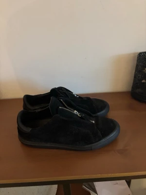 Svarta axel arigato sneakers med dragkedja - Svarta sneakers i mocka med dragkedja på ovansidan istället för klassiska snören. Snygg och stilren design med låg profil och svart sula. Perfekta för dig som gillar en clean look och vill ha något enkelt men ändå trendigt.