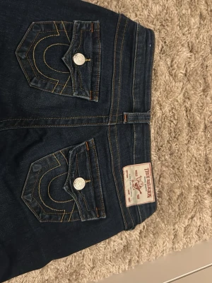 True Religion mörkblå jeans - Säljer ett par mörkblå jeans från True Religion med ikoniska kontrastsömmar och snygga fickdetaljer med silverknappar. Jeansen har flare-modell och är tillverkade i kraftigt denimtyg. Väldigt stretchigt material så passar mig som har storlek 27💗