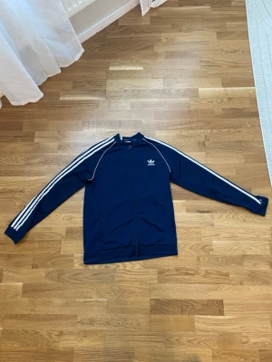 Marinblå Adidas track jacket - Snygg marinblå track jacket från Adidas med klassiska vita ränder längs ärmarna och vit logga på bröstet. Dragkedja framtill och hög krage. Tillverkad i ett mjukt och stretchigt material som ger en sportig look.