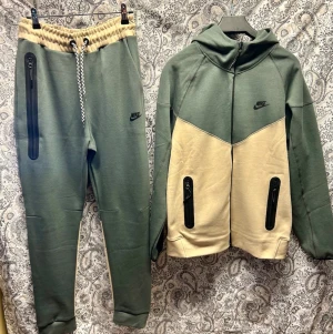 Nike loungewear set i grönt och beige - Nike loungewear set med hoodie och byxor i olivgrönt och beige. Hoodien har dragkedja, huva och svarta detaljer med fickor framtill. Byxorna har dragsko i midjan och svarta dragkedjefickor. Perfekt för chill dagar hemma.