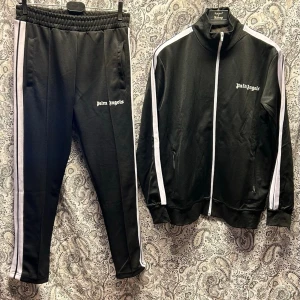 Svart tracksuit set från Palm Angels - Snyggt svart tracksuit set från Palm Angels med vita ränder längs ärmar och ben. Jackan har dragkedja, hög krage och två fickor framtill. Byxorna har resår i midjan och raka ben med vita sidolinjer. Loggan är tryckt i vitt på både jacka och byxa.
