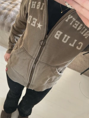 Grön Ed Hardy zip-hoodie - Grön hoodie från Ed Hardy med dragkedja, huva med dragsko och stora fickor fram. Tryck i vitt på bröstet och ärmen ger en cool streetkänsla. Perfekt för dig som gillar avslappnad stil och vill ha något bekvämt och snyggt.