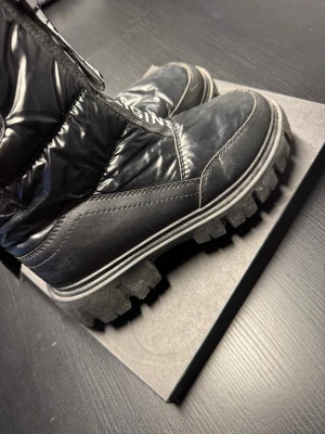 Svarta moon boots från Different - Säljer ett par svarta boots med chunky sula och glansig ovandel. Bootsens baksida har en logga och dragkedjedetalj. Materialet är en mix av syntet och textil, perfekt för dig som vill sticka ut med en cool vinterlook.