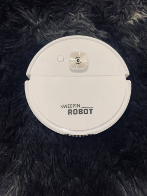 Sweepin Robot vit robotdammsugare - Säljer en vit robotdammsugare från Sweepin Robot. Den har rund form, en stor silverfärgad knapp på ovansidan och är tillverkad i plast. Perfekt för att automatiskt hålla golvet rent hemma utan ansträngning.