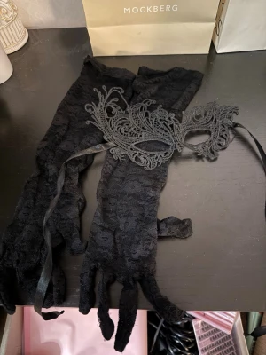 Svart spetsmask och handskar - Säljer en svart maskeradmask i spets med matchande långa handskar. Masken har fina broderade detaljer och knyts med band. Handskarna är i genombruten spets och går upp över armbågen. Perfekt för maskerad eller cosplay.