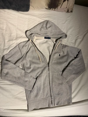 Polo Ralph Lauren Zip - Snygg grå ziphoodie med dragkedja från Polo Ralph Lauren. Klassisk design med huva och vita snören, samt broderad logga på bröstet. Mjuk insida och två fickor framtill. Perfekt för en avslappnad och stilren look. Välldigt bra skick 10/10, inga skador eller likande alls. Säljer då den tyvärr blivit för liten. 