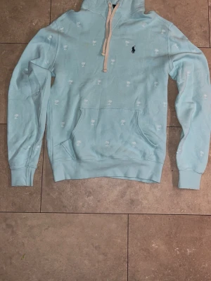 Ljusblå hoodie från Polo Ralph Lauren - Säljer en ljusblå hoodie från Polo Ralph Lauren med broderade vita palmträd och klassiska mörkblå loggan på bröstet. Hoodien har huva med beige dragsko och en stor magficka. Perfekt för dig som gillar stilren men ändå chill look.