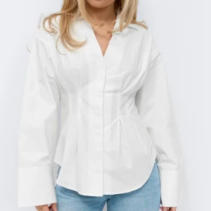 Vit figursydd blus med v-ringning - Stilren vit blus med markerad midja och v-ringad hals. Blusen har långa ärmar med breda manschetter och en slät framsida utan synliga knappar. Perfekt för en trendig och clean look. Slutsåld på ginas hemsida