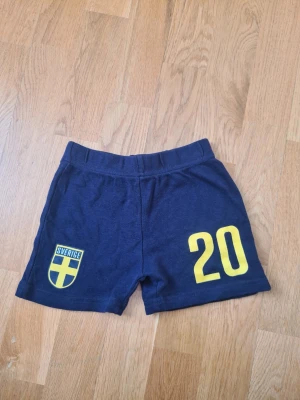 Blå Sverige shorts med nummer 20 - Mörkblå shorts med gult Sverige-emblem och siffran 20 framtill. Perfekta för fotboll eller träning, med elastisk midja för skön passform. Tillverkade i mjuk bomull som andas. Storlek 74/80.