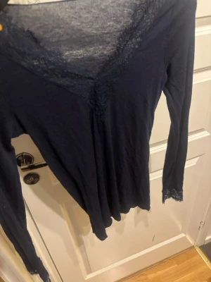 Mörkblå långärmad topp med spets - Mörkblå långärmad topp med v-ringning och spetsdetaljer längs halsringningen och ärmsluten. Tunn och mjuk i materialet, perfekt för lager på lager eller som den är. Enkel men ändå elegant design.Storlek S men sitter som Xs