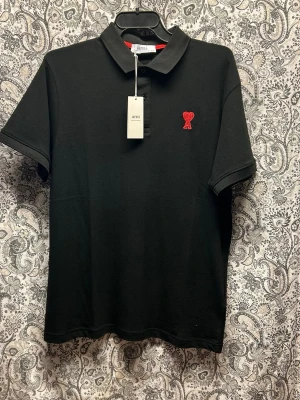 Svart pikétröja från Ami Paris - Snygg svart pikétröja från Ami Paris med klassisk krage och knappar framtill. Tröjan har ett rött broderat hjärta med Ami-loggan på bröstet. Tillverkad i mjuk bomull och har korta ärmar. Perfekt för en clean och stilren look.